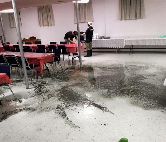 The inside of a cafeteria in a church, with a large, dirty puddle in the middle of the floor. Two staff at the end.
